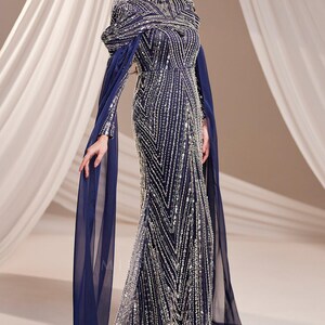 May include: A navy blue evening gown with intricate silver beadwork. The dress features a high neckline, long sleeves, and flowing cape-like sleeves. The gown's design includes vertical and chevron patterns, creating a glamorous and elegant look.