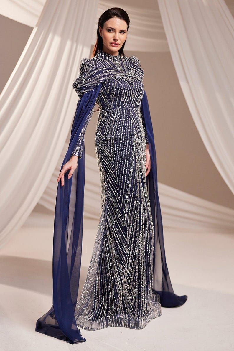 May include: A navy blue evening gown with intricate silver beadwork. The dress features a high neckline, long sleeves, and flowing cape-like sleeves. The gown's design includes vertical and chevron patterns, creating a glamorous and elegant look.