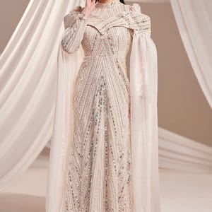 May include: A floor-length, cream-colored gown with intricate beadwork and a high neckline. The dress features long sleeves and sheer cape-like fabric extending from the shoulders. The gown's design includes vertical and diagonal beaded patterns.