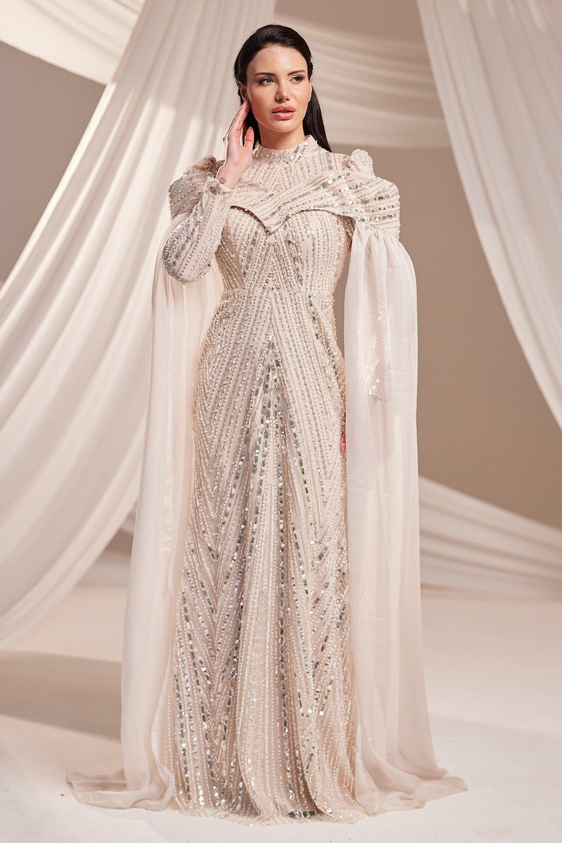 May include: A floor-length, cream-colored gown with intricate beadwork and a high neckline. The dress features long sleeves and sheer cape-like fabric extending from the shoulders. The gown's design includes vertical and diagonal beaded patterns.