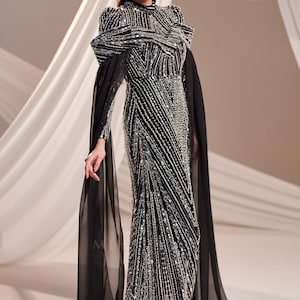 May include: A black and silver beaded evening gown with a high neckline and long sleeves. The dress features a fitted silhouette with a flowing skirt and sheer black cape sleeves. The gown is embellished with intricate beadwork.