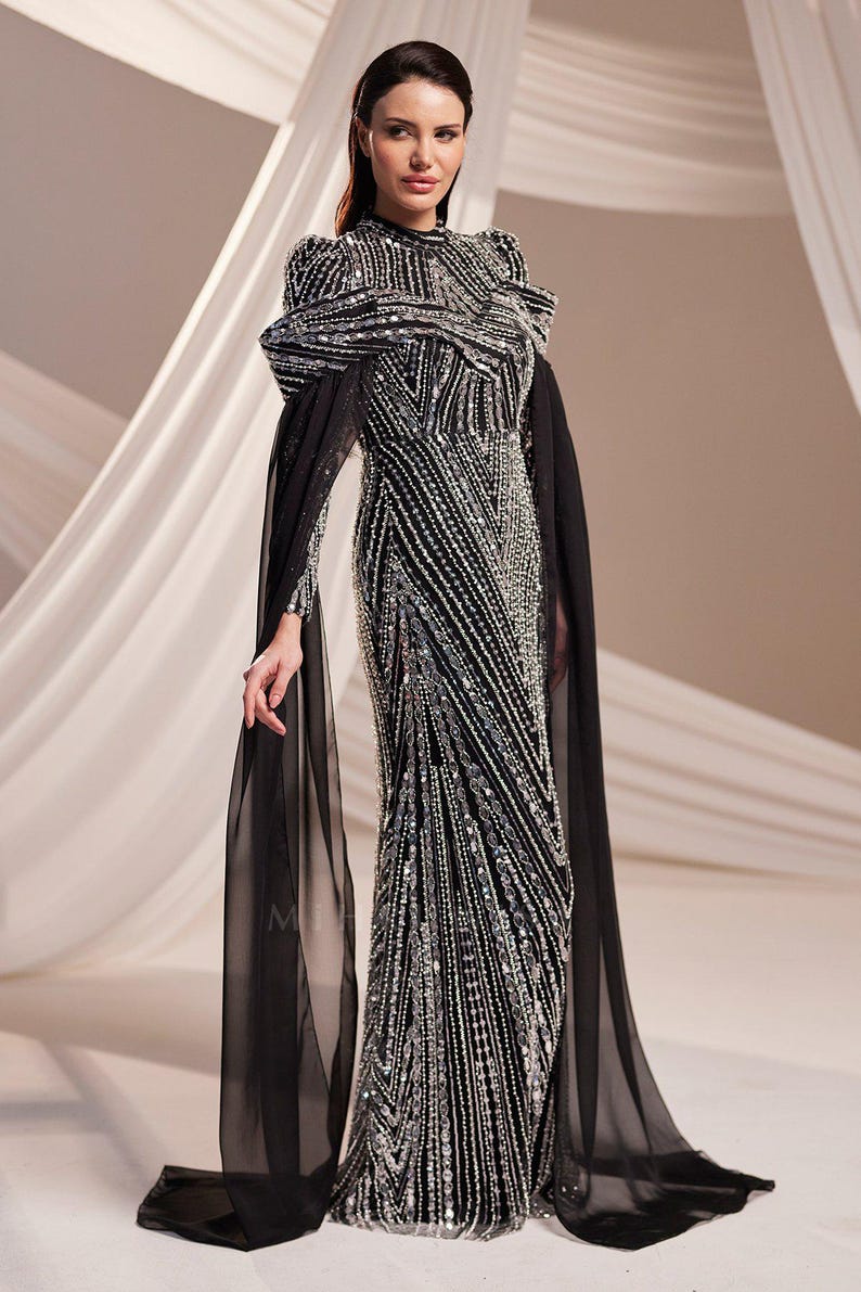 May include: A black and silver beaded evening gown with a high neckline and long sleeves. The dress features a fitted silhouette with a flowing skirt and sheer black cape sleeves. The gown is embellished with intricate beadwork.