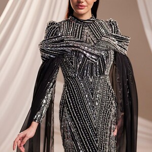 May include: A black evening gown with silver sequin and bead embellishments. The dress features a high neckline, long sleeves, and a flowing skirt. The shoulders have a unique design, and sheer black fabric drapes from the sleeves.