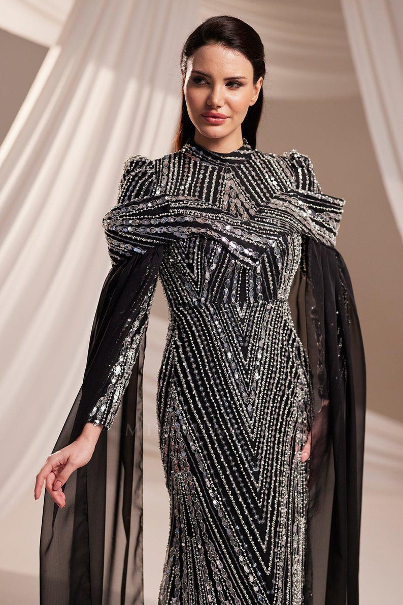 May include: A black evening gown with silver sequin and bead embellishments. The dress features a high neckline, long sleeves, and a flowing skirt. The shoulders have a unique design, and sheer black fabric drapes from the sleeves.