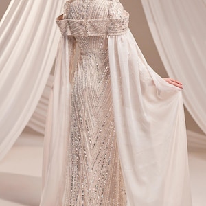 May include: A floor-length, ivory-colored gown with intricate beadwork and a high neckline. The dress features long, flowing sleeves and a cape-like overlay. The gown is embellished with silver sequins, creating a shimmering effect.