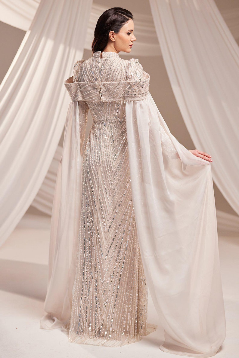 May include: A floor-length, ivory-colored gown with intricate beadwork and a high neckline. The dress features long, flowing sleeves and a cape-like overlay. The gown is embellished with silver sequins, creating a shimmering effect.