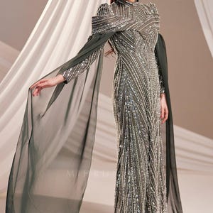 May include: An olive green formal gown with intricate silver beading and a high neckline. The dress features long, sheer cape sleeves that drape elegantly. The gown is floor-length and suitable for special occasions.