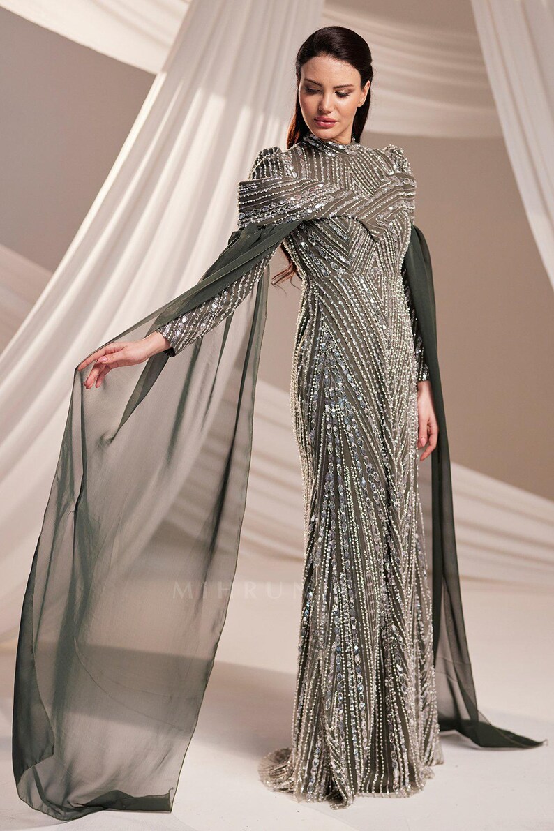 May include: An olive green formal gown with intricate silver beading and a high neckline. The dress features long, sheer cape sleeves that drape elegantly. The gown is floor-length and suitable for special occasions.