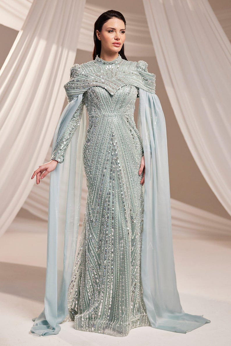 May include: A light green, floor-length gown with intricate beadwork and a high neckline. The dress features long, flowing sleeves and a cape-like overlay in a matching light blue hue. The gown is embellished with sparkling sequins.