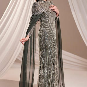 May include: A formal, floor-length gown with intricate beadwork and a high neckline. The dress is a muted green color with silver embellishments. Long, flowing cape sleeves extend from the shoulders. The dress is suitable for special occasions.