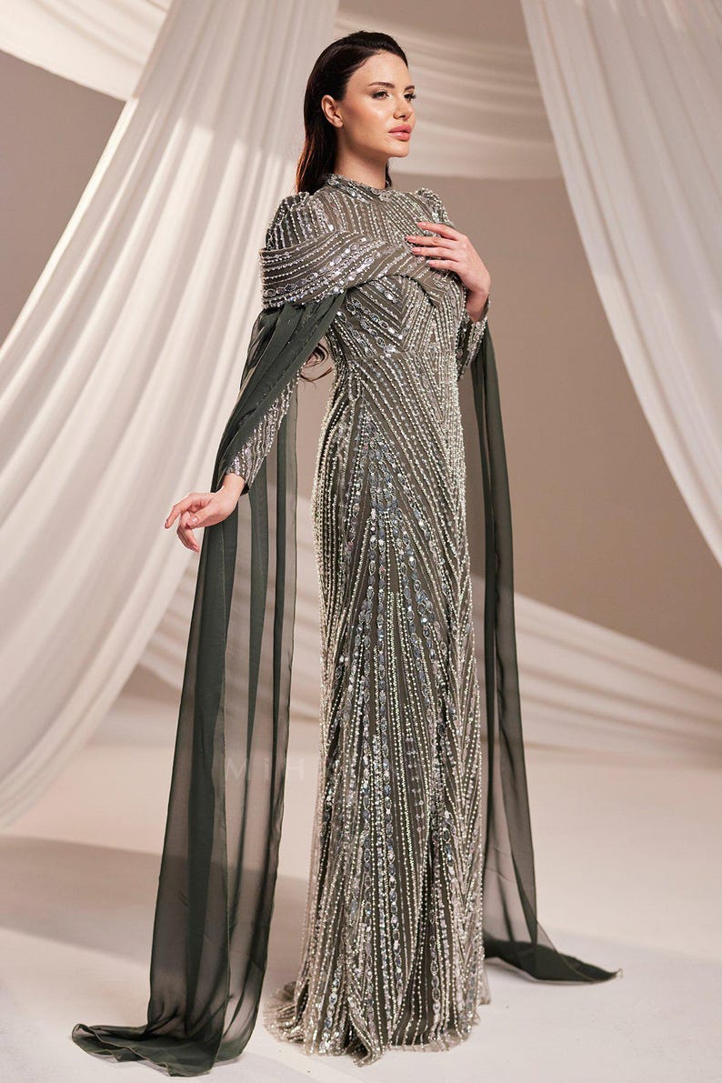 May include: A formal, floor-length gown with intricate beadwork and a high neckline. The dress is a muted green color with silver embellishments. Long, flowing cape sleeves extend from the shoulders. The dress is suitable for special occasions.