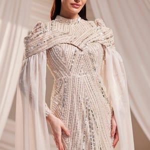 May include: Elegant ivory-colored gown with intricate beadwork and sequin embellishments. Features a high neckline, long sleeves, and a flowing cape-like overlay. The dress is floor-length, suitable for formal events.