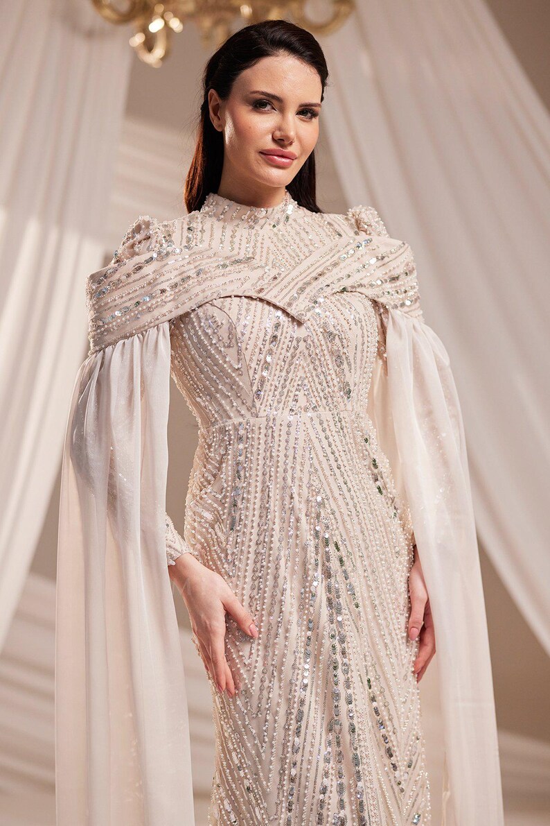 May include: Elegant ivory-colored gown with intricate beadwork and sequin embellishments. Features a high neckline, long sleeves, and a flowing cape-like overlay. The dress is floor-length, suitable for formal events.