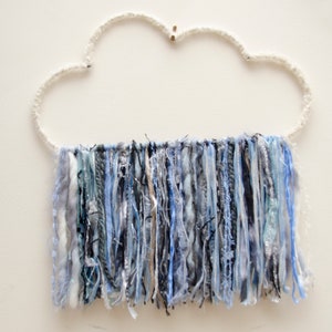 May include: A cloud-shaped wall hanging with a white, textured frame and dangling strands of blue, silver, and gray yarn. The yarn strands create a textured, layered effect, adding visual interest to the piece.
