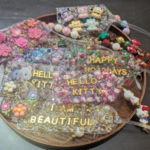 May include: A collection of decorative resin tags with various embellishments. Some tags feature Hello Kitty characters, flowers, and the phrases "Hello Kitty", "Happy Holidays", and "I Am Beautiful". The tags are adorned with beads and glitter.