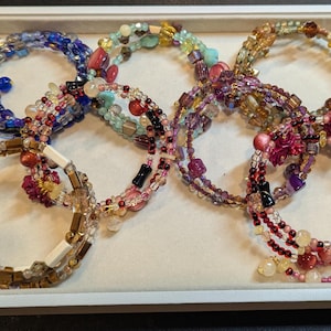 Puede incluir: Una colección de pulseras de cuentas en varios colores, incluyendo azul, rosa, dorado y morado. Las pulseras están dispuestas en una bandeja blanca, mostrando sus intrincados diseños y texturas. Cada pulsera tiene un diseño único.