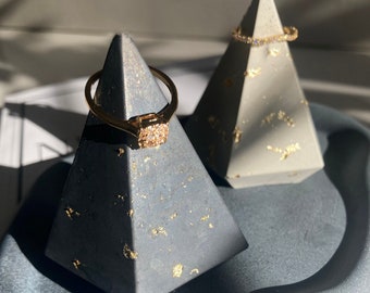 May include: Two gray concrete pyramid ring holders with gold flecks. One holder has a gold ring with a square-shaped stone on it. The other holder has a gold ring with small stones around it. The holders are on a gray, textured surface.