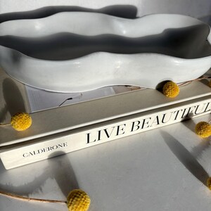 May include: A white ceramic wavy dish with a rough texture sits on top of a book titled "Live Beautiful" by Calderone.  Dried yellow flowers are scattered around the dish.