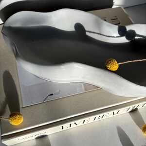 May include: A white ceramic wavy dish with a book titled "Live Beautiful" by Calderoni. The book is on a white surface with yellow dried flowers.
