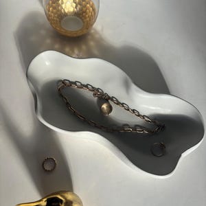 May include: A white ceramic dish with a wavy edge, featuring a gold chain necklace and two gold rings. A gold bird figurine is in the foreground.
