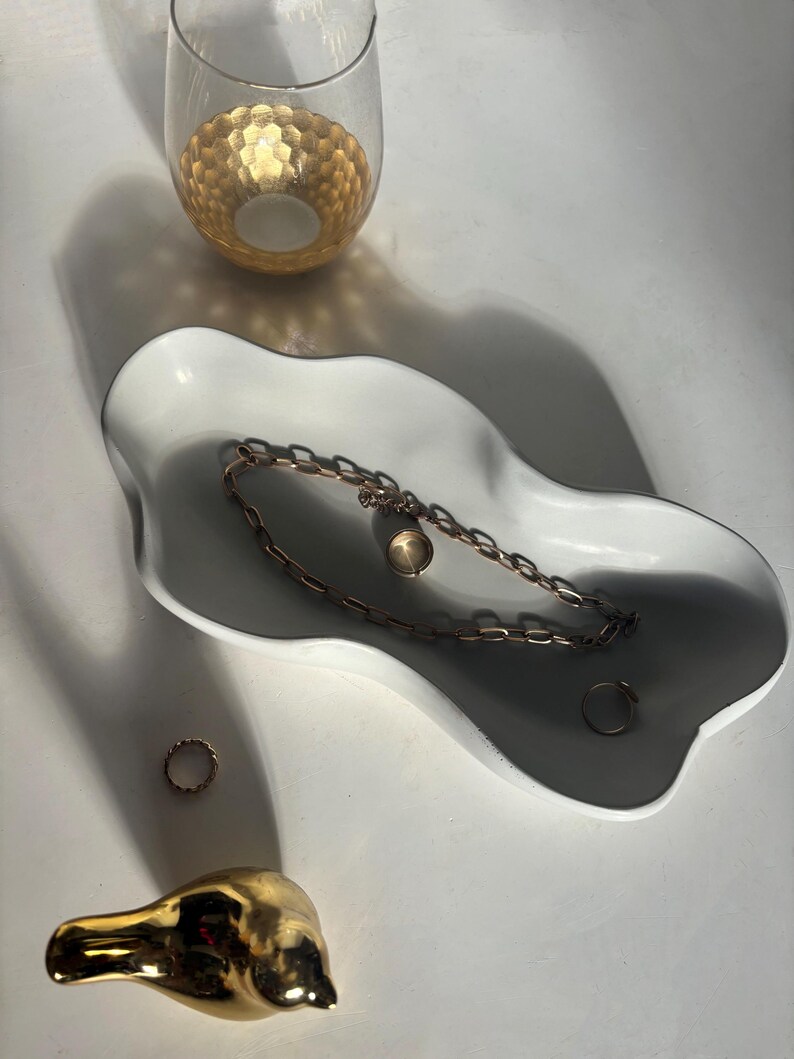 May include: A white ceramic dish with a wavy edge, featuring a gold chain necklace and two gold rings. A gold bird figurine is in the foreground.