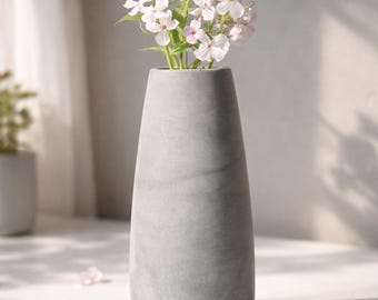Può includere: Un vaso grigio cilindrico contiene un bouquet di fiori bianchi e rosa. Il vaso ha una finitura liscia e opaca e una base leggermente arrotondata. Petali sparsi degli stessi fiori sono sulla superficie. Il vaso è un oggetto decorativo per la casa.