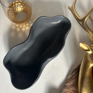 May include: A black, wavy, ceramic bowl with a smooth, glossy finish. The bowl is shaped like a long, narrow oval with a slight curve in the middle.