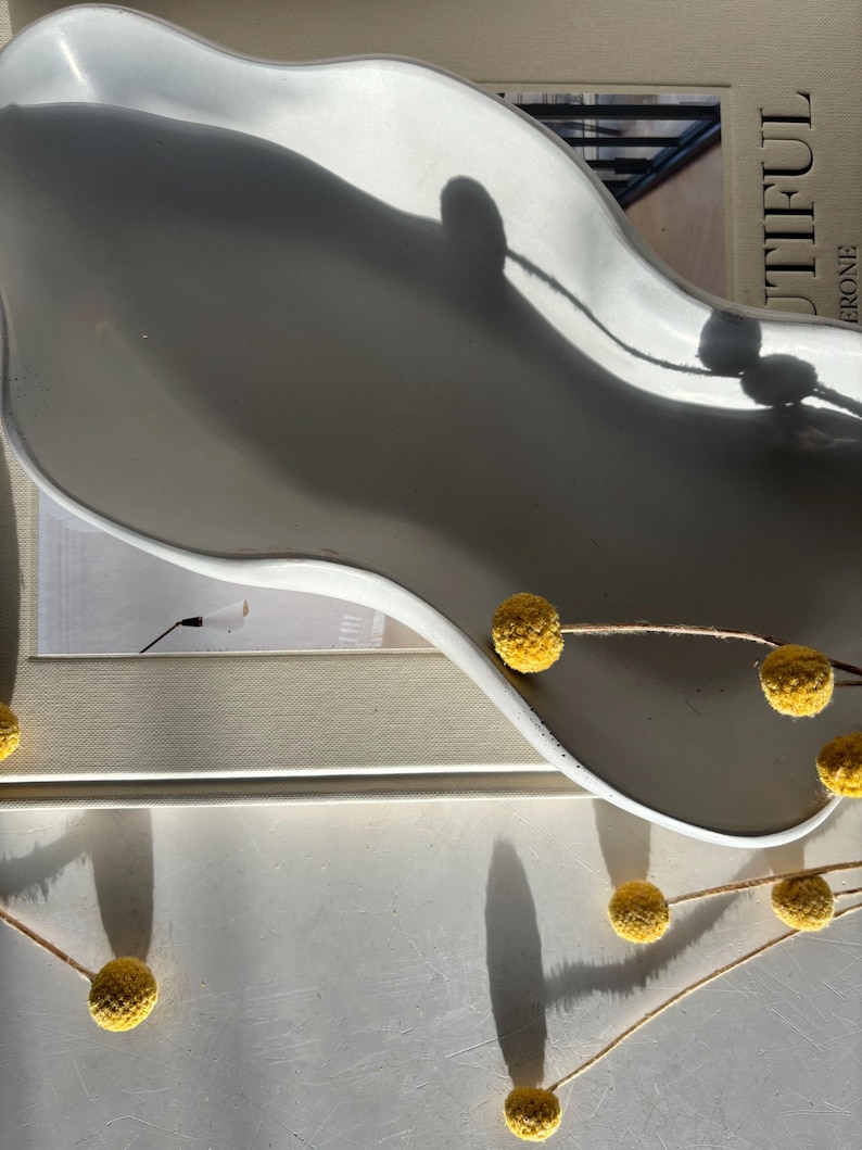 May include: White ceramic tray with a wavy edge, featuring a shadow of a window. The tray is decorated with a string of small yellow dried flowers.