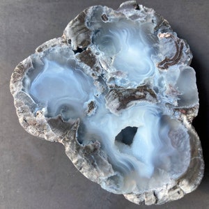 Puede incluir: Una geoda con un interior blanco translúcido y un exterior gris rugoso. La geoda tiene una forma natural y orgánica y presenta varias cavidades.