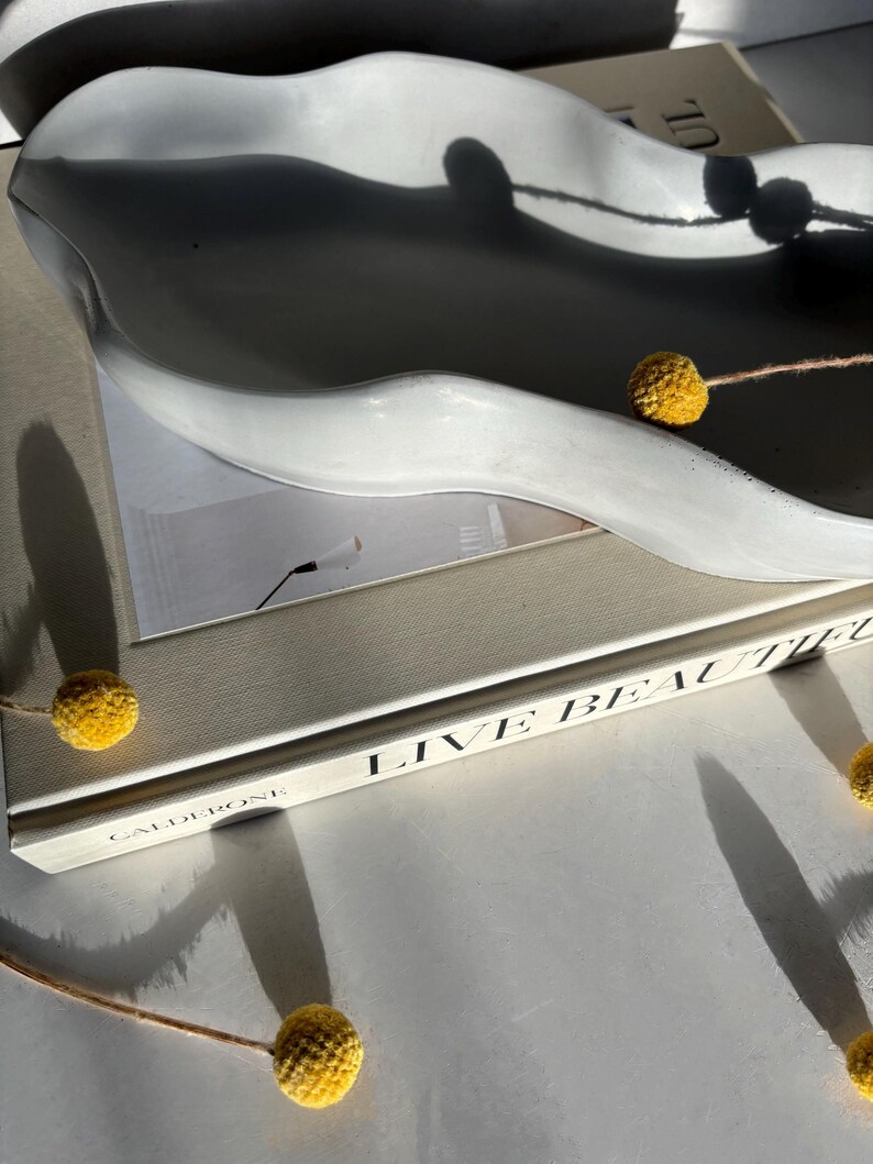 May include: A white ceramic wavy dish with a book titled "Live Beautiful" by Calderoni. The book is on a white surface with yellow dried flowers.
