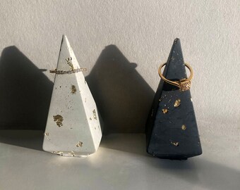 May include: Two geometric ring holders. One is white with gold leaf accents, the other is black with gold leaf accents. A gold ring with small stones sits on the white holder, and a gold ring with a square stone sits on the black holder.