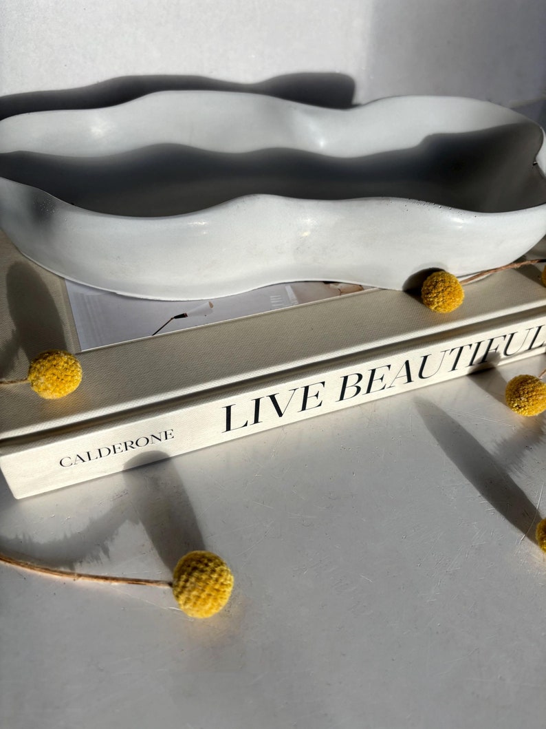 May include: A white ceramic wavy dish with a rough texture sits on top of a book titled "Live Beautiful" by Calderone.  Dried yellow flowers are scattered around the dish.