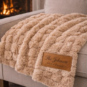 Può includere: Una morbida coperta color beige con un motivo intrecciato e strutturato è drappeggiata su un divano. Una toppa rettangolare in pelle con la scritta "The Johnson Family" è cucita sulla coperta. Sullo sfondo si vede un camino.