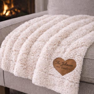 Può includere: Coperta in peluche color crema con una trama a coste, drappeggiata su un divano grigio. Una toppa in pelle a forma di cuore con la scritta "The Johnson Family" è cucita sulla coperta. Un camino è visibile sullo sfondo.