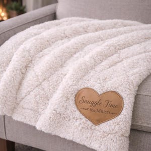 Może przedstawiać: Kremowy, pluszowy koc z sercowatą skórzaną naszywką z napisem "Snuggle Time" i nazwiskiem. Koc jest narzucony na szarą sofę, tworząc przytulną i zachęcającą scenę.