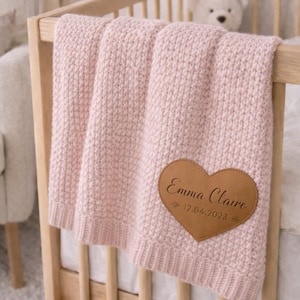 Può includere: Una coperta per neonati rosa chiaro lavorata a maglia, drappeggiata su una culla di legno. La coperta presenta una toppa in pelle a forma di cuore con il nome "Emma Claire" e la data "12.04.2023" incisi. Un orsacchiotto bianco è visibile.