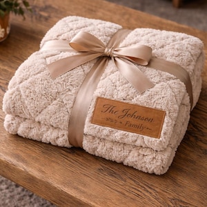 Può includere: Una coperta beige e soffice con un motivo a rombi, legata con un nastro e un fiocco di raso. Una toppa in pelle marrone mostra il testo "The Johnson Family". La coperta è piegata e presentata su una superficie di legno.