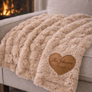 Può includere: Una soffice coperta beige con una toppa in pelle a forma di cuore. La toppa recita "The Johnson Family". La coperta è drappeggiata su un divano grigio, con un camino sullo sfondo.