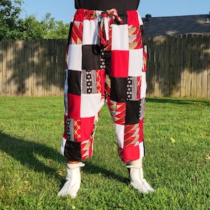 May include: Patchwork trousers in red, black, and white squares. The trousers have a drawstring waist and are cropped at the calf. The fabric features various patterns, including stripes and geometric designs. The trousers are worn with white boots.