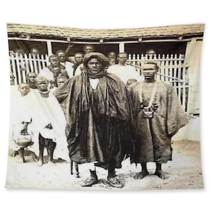 May include: A group of people, including men and children, stand in front of a building. The men are wearing traditional clothing, and the children are holding bowls. The image is in black and white.