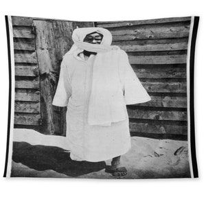 May include: A black and white photograph of a person wearing a white robe and a white head covering. The person is standing in front of a wooden wall.
