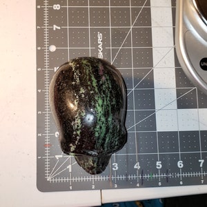 Puede incluir: Un cr&aacute;neo de piedra verde, negro y rosa tallado en una piedra pulida. El cr&aacute;neo est&aacute; sobre una estera de corte gris con marcas blancas.