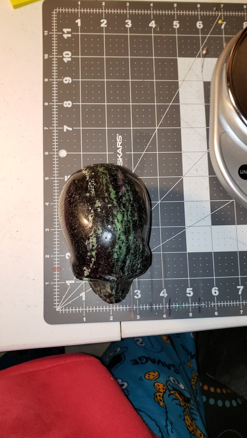 Puede incluir: Un cr&aacute;neo de piedra verde, negro y rosa tallado en una piedra pulida. El cr&aacute;neo est&aacute; sobre una estera de corte gris con marcas blancas.