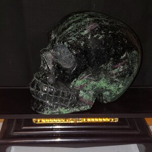 Puede incluir: Un cr&aacute;neo de piedra negra y verde tallado en una sola pieza de piedra. El cr&aacute;neo est&aacute; sentado sobre un soporte de madera negro.