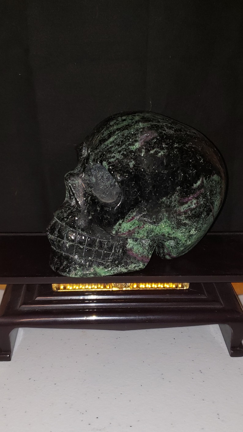 Puede incluir: Un cr&aacute;neo de piedra negra y verde tallado en una sola pieza de piedra. El cr&aacute;neo est&aacute; sentado sobre un soporte de madera negro.