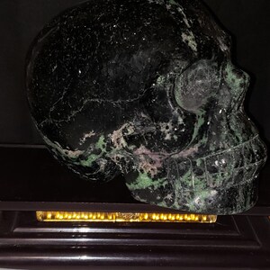 Puede incluir: Un cr&aacute;neo de cristal negro y verde con una talla detallada. El cr&aacute;neo est&aacute; sentado sobre una base de madera negra con una tira de luz dorada.