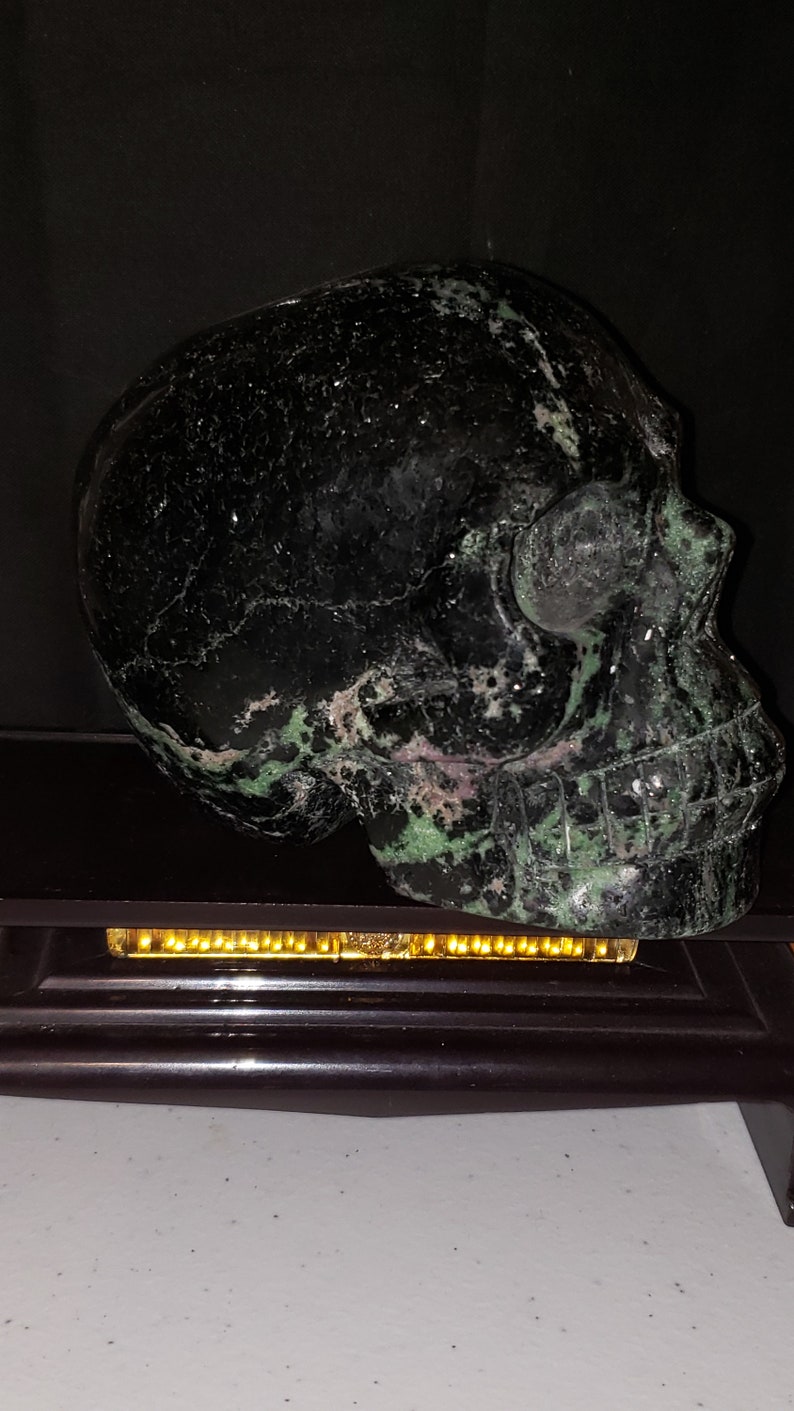 Puede incluir: Un cr&aacute;neo de cristal negro y verde con una talla detallada. El cr&aacute;neo est&aacute; sentado sobre una base de madera negra con una tira de luz dorada.