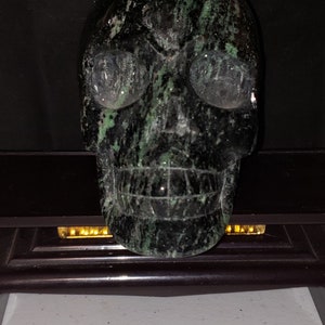 Pode incluir: Um crânio preto e verde feito de pedra. O crânio tem uma escultura detalhada dos traços faciais e um padrão de mármore verde e preto.