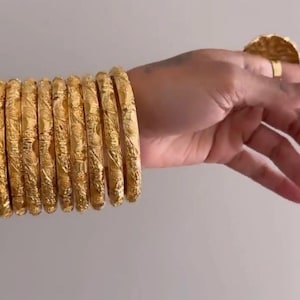 Puede incluir: Una colección de pulseras doradas y un anillo. Las pulseras son gruesas, con intrincados diseños en relieve. El anillo tiene un gran diseño circular texturizado. Las joyas se exhiben sobre un fondo neutro.
