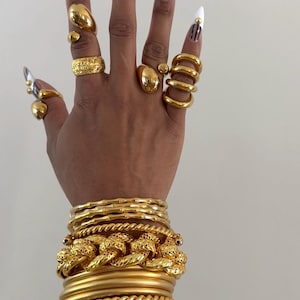 Puede incluir: Una mano adornada con joyas de oro, incluyendo múltiples anillos de varios diseños y una pila de brazaletes dorados. Las uñas largas están pintadas con un diseño de rayas de cebra y blanco.